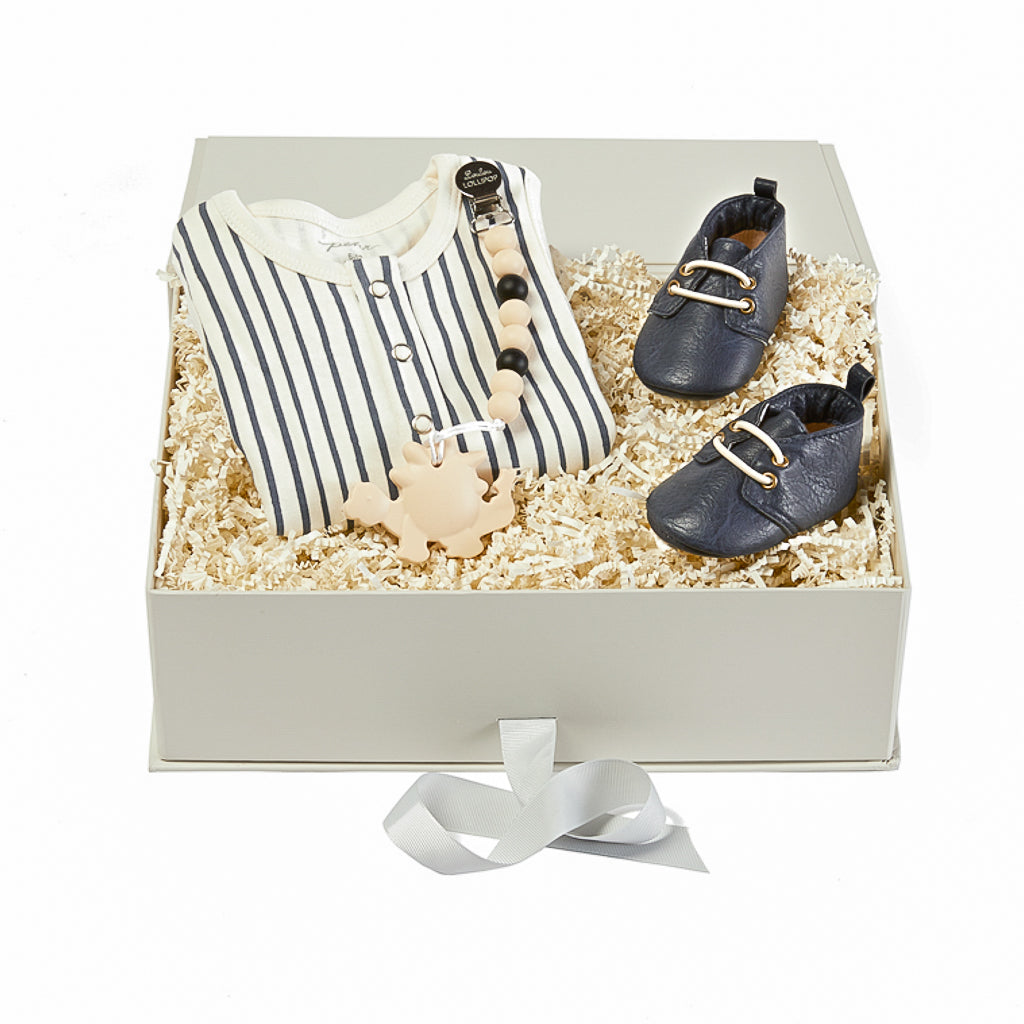 anna and amy gift box for boys, including navy blue stripe onesie, navy blue baby shoes, and a cream dinosaur teether.